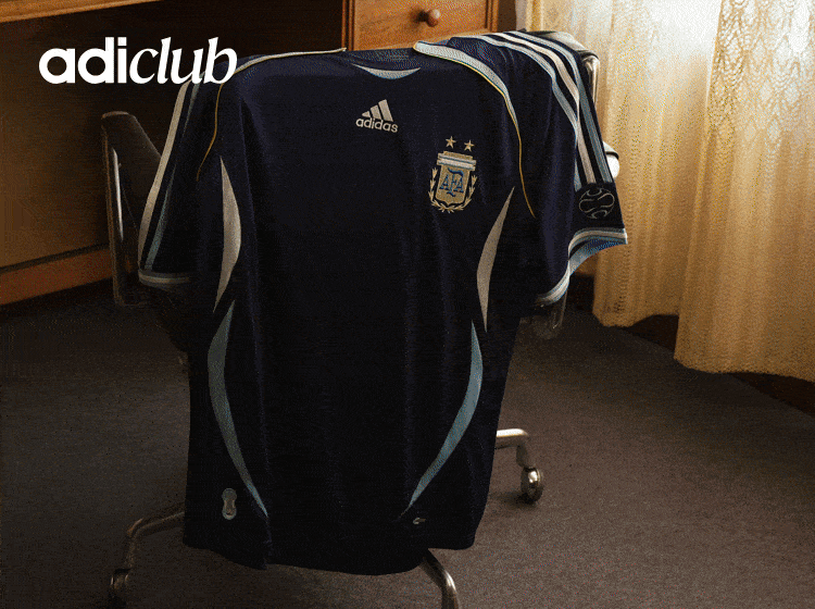 The image shows a black and blue Adidas soccer jersey with the Adidas logo and the Argentine national team crest prominently displayed. The jersey is hanging on a wooden rack in what appears to be a room with curtains in the background.