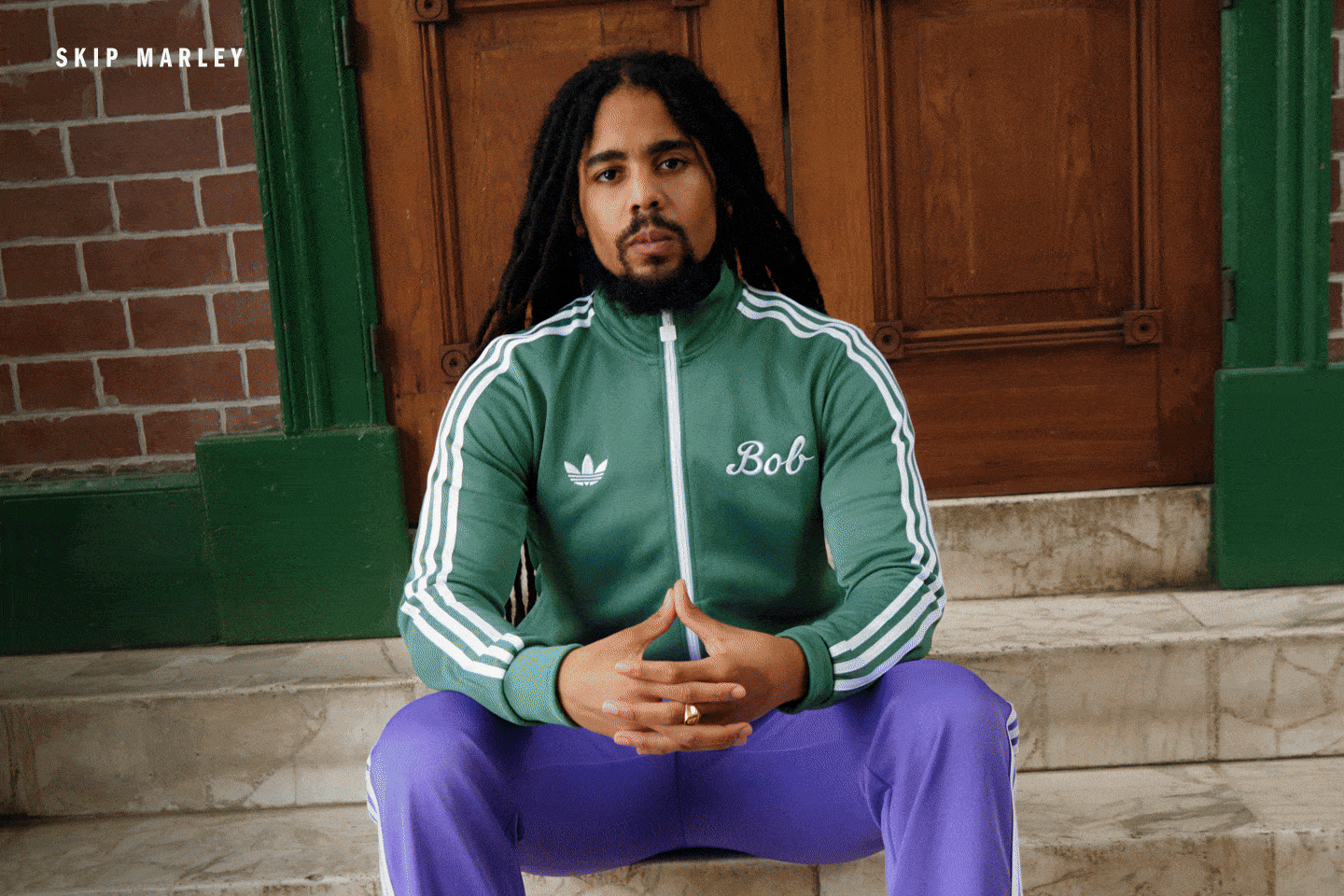 The image shows a person with long dark hair wearing a green jacket with white stripes, sitting on a brick wall in front of a green door.