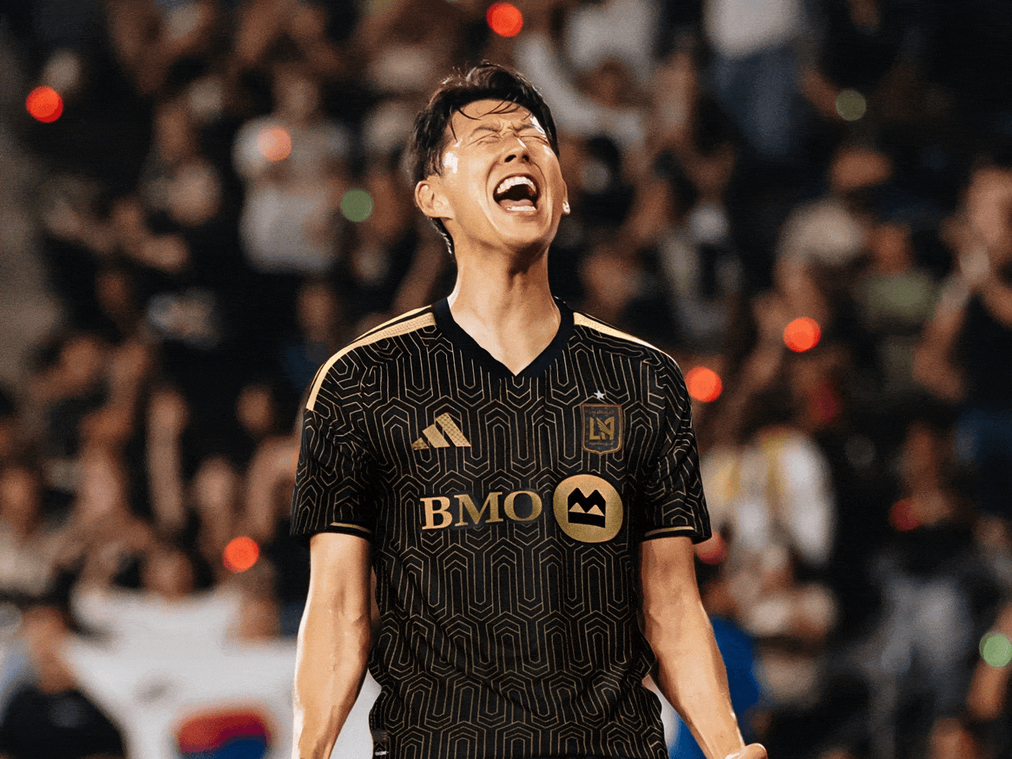 A person in a black and gold soccer jersey is celebrating with an exuberant expression on a dimly lit field surrounded by a crowd.