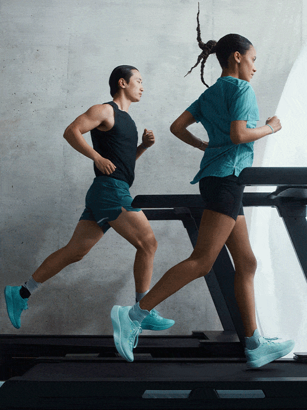 Two people, a man and a woman, are running on treadmills in a dimly lit gym setting with a concrete wall in the background.