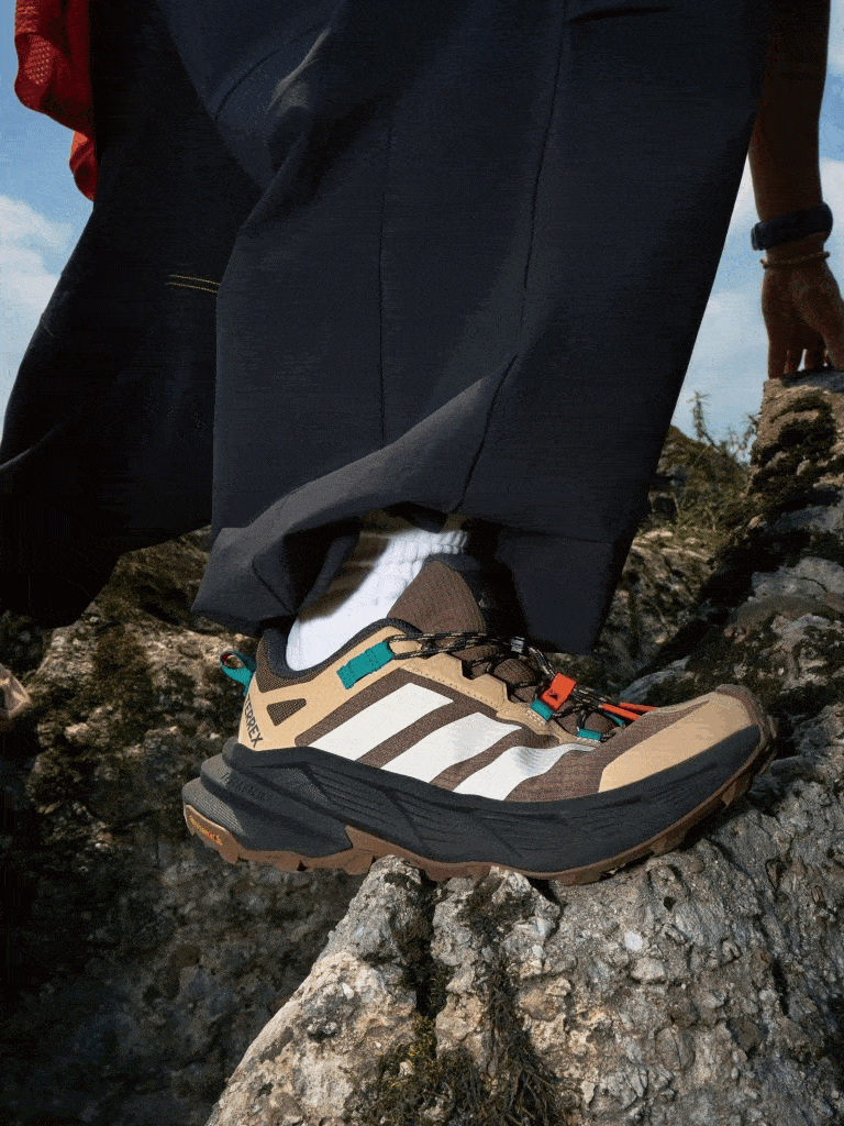 The image shows a person's lower body wearing dark pants and hiking boots standing on a rocky, mountainous terrain with a cloudy sky in the background.