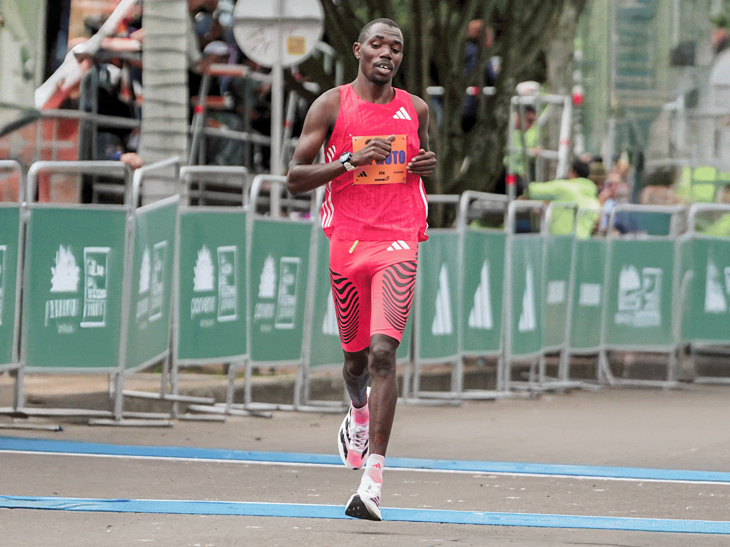 A male athlete in a red and white athletic uniform is running on a track surrounded by barriers and spectators in the background.