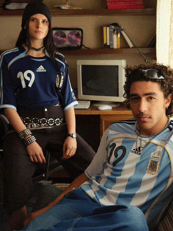 The image shows two young people, a woman and a man, sitting in a room with shelves and a computer monitor in the background.
