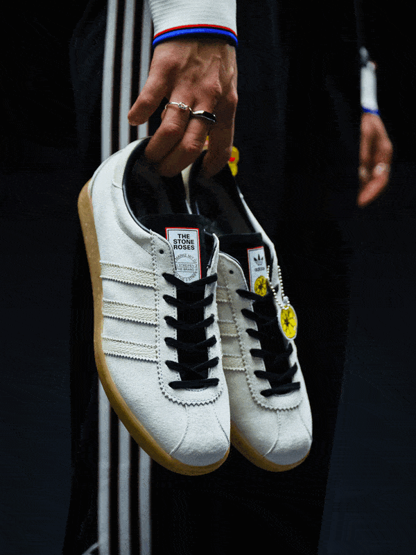 A pair of white and black sneakers with yellow accents are being held up against a dark background, showcasing the intricate design and details of the footwear.