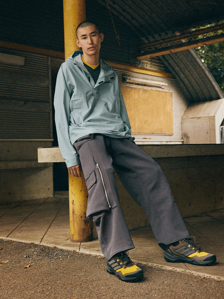 A young person wearing a gray jacket and dark pants is sitting on a wooden structure in what appears to be a rural or outdoor setting.