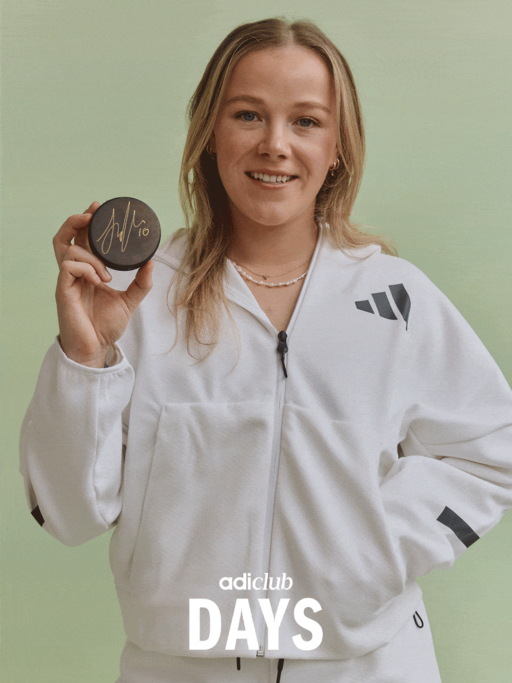 A smiling young woman in a white athletic jacket holds a small round object, likely a medal or award, against a light green background.