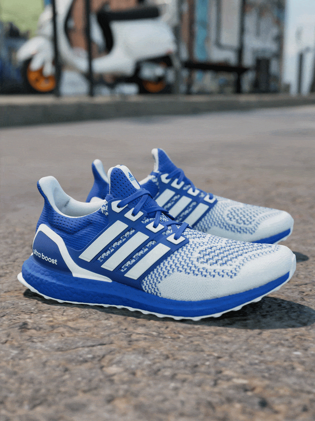 A pair of blue and white Adidas Ultra Boost running shoes sits on a concrete surface, with a blurred urban background in the distance.