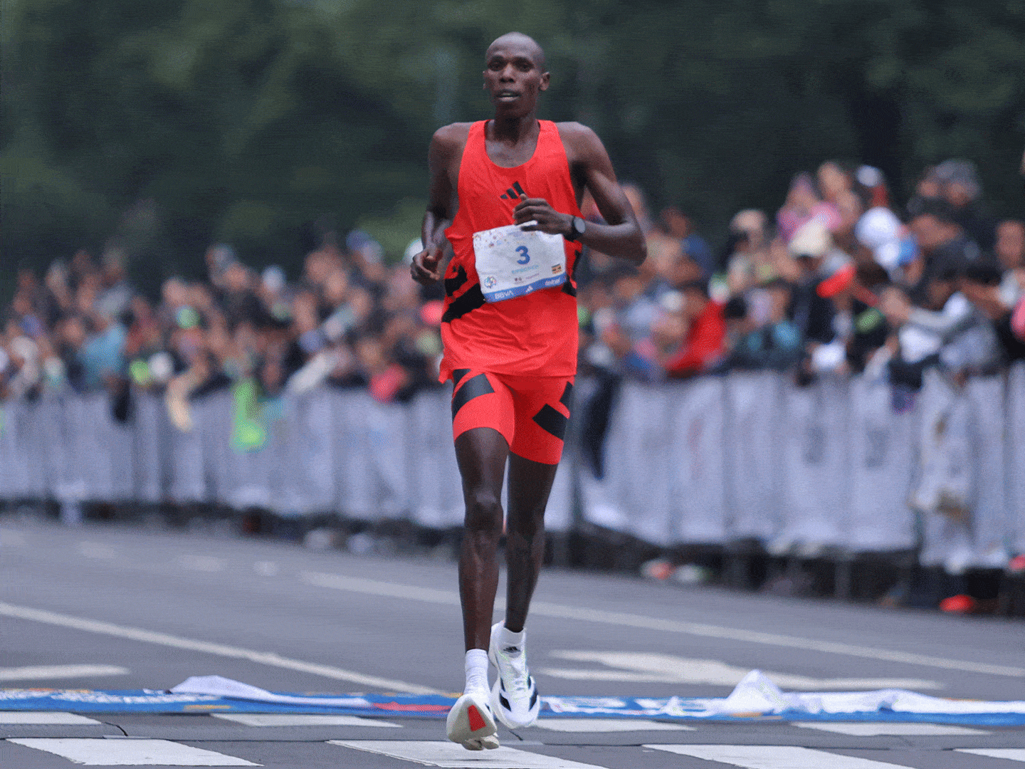 A male runner in a red uniform is running on a road surrounded by spectators and barriers, with a blurred background of trees and buildings.