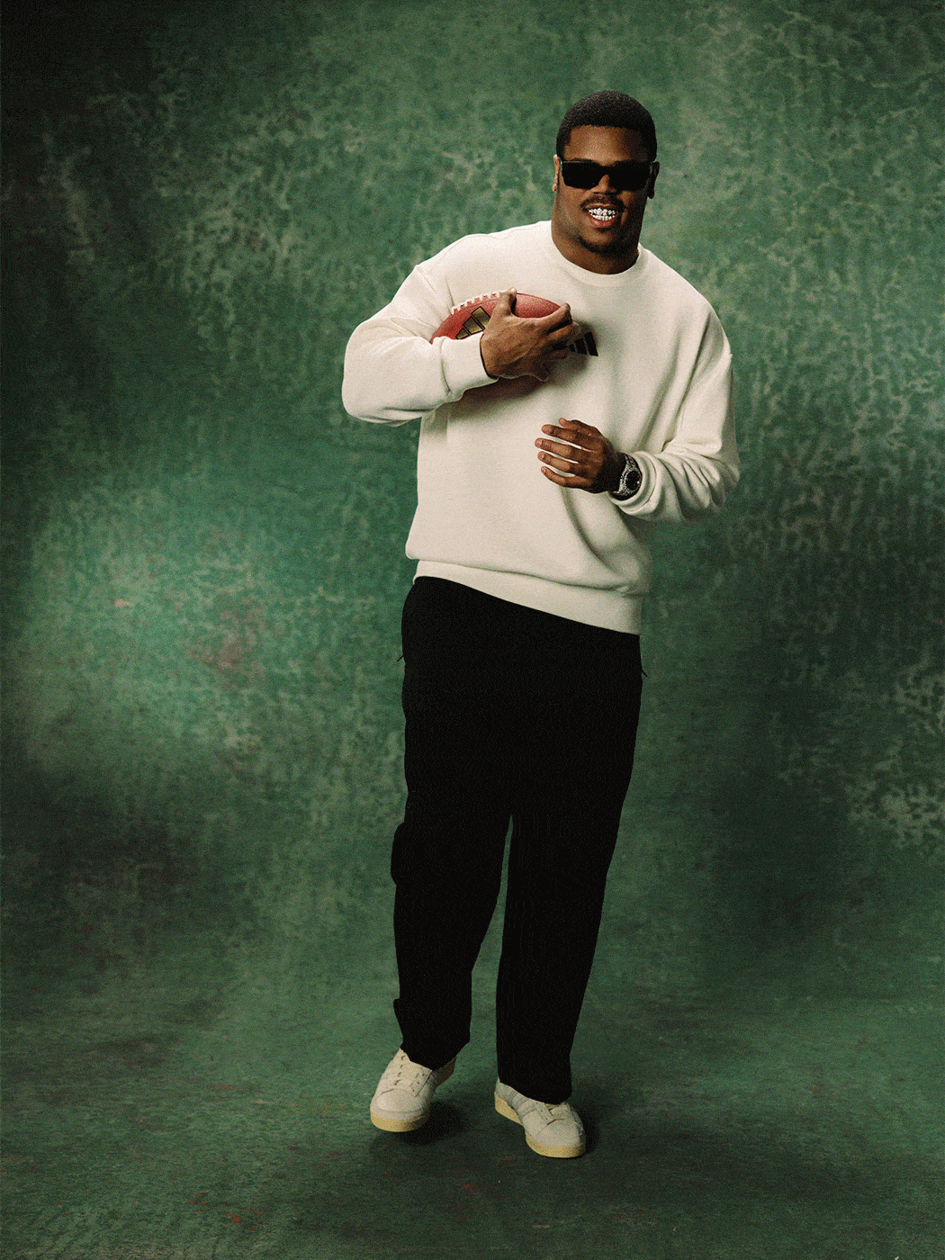 A person wearing a white sweater and black pants stands in front of a green background, appearing to be in a dance or performance pose.