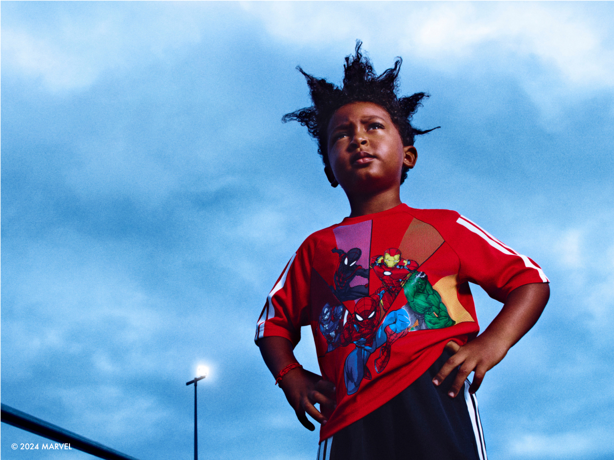 A young person wearing a vibrant red shirt with colorful designs stands against a cloudy blue sky backdrop.