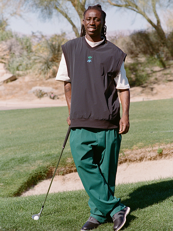 A man wearing a gray shirt and green pants is standing on a grassy field, holding a golf club, with trees and a clear sky visible in the background.