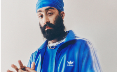 A portrait of a man in a blue turban and a blue adicolor tracksuit standing in front of a white backdrop. 