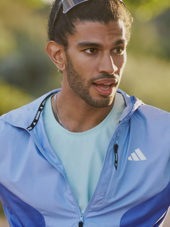 A man with brown hair wears a light blue adidas tracksuit and a turquoise t-shirt while jogging in nature. 