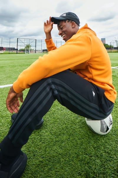 English football player Noni Madueke sits on a football pitch in adidas clothing, smiling as he adjusts his cap.