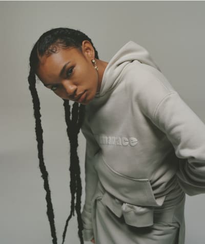 Model leans forward looking to camera wearing gray hoodie against neutral gray background.