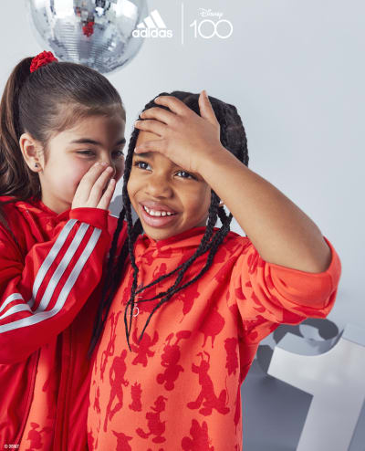 Two children, one whispering in the others ear as the other covers her mouth, both wearing the adidas | Disney100 collection.