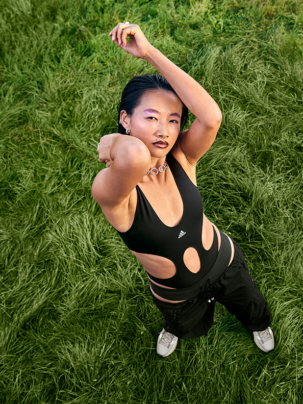 Asian female model wears a black adidas bodysuit with cutouts exposing her midriff and track pants sitting low on her hips.