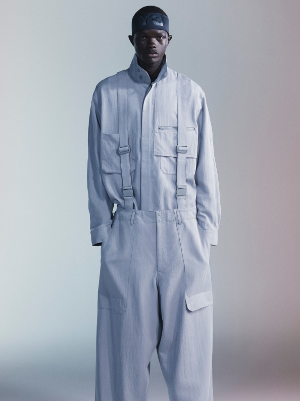 A man in a gray adidas Y-3 overall stands casually with his hands in his pockets against a white background.