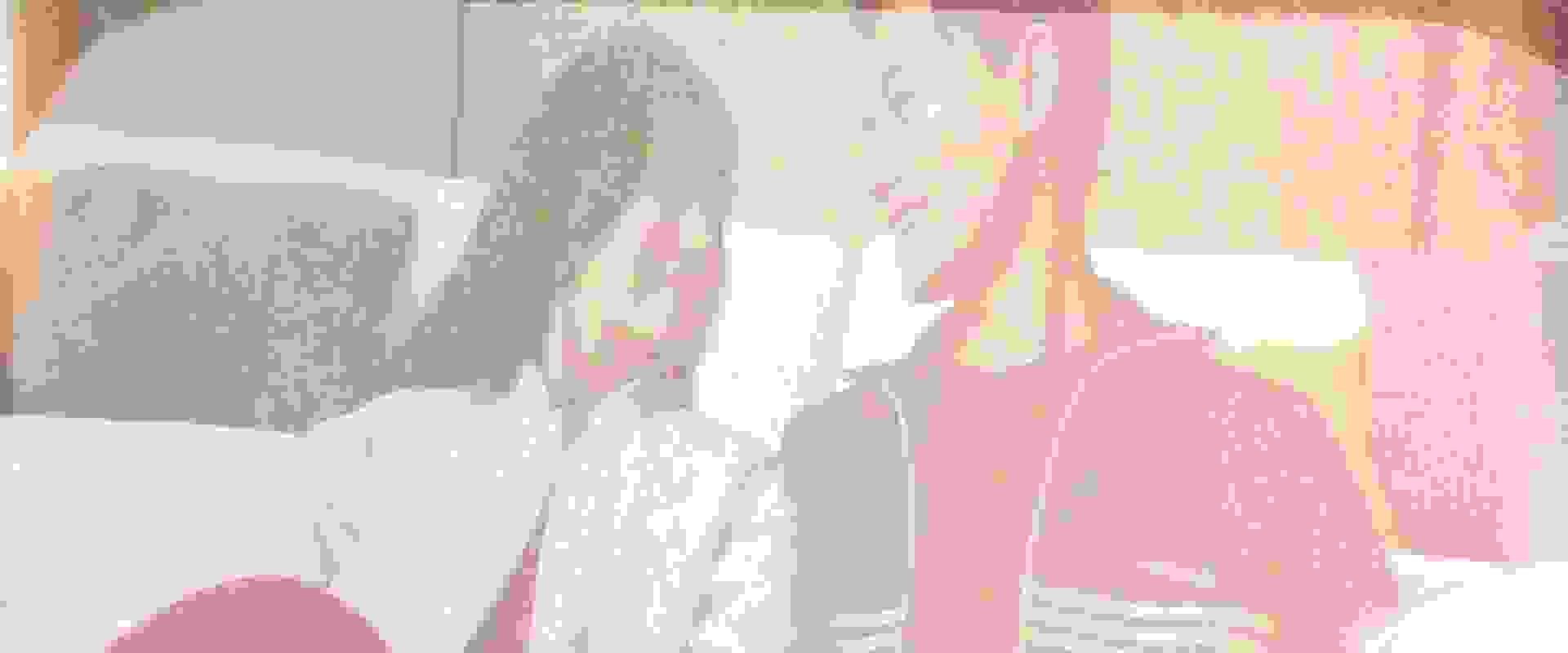 Two individuals, a man and a woman, are seated on a couch in a room with a bookshelf visible in the background.