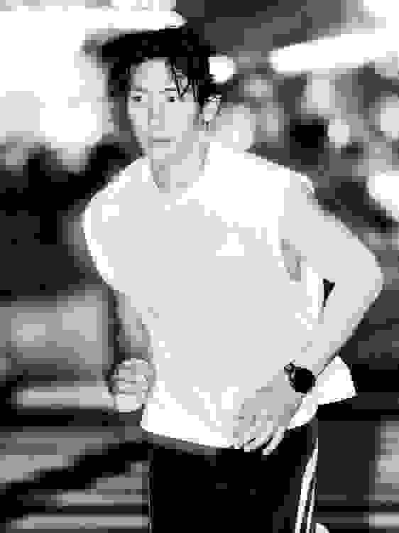 A person wearing a white shirt and black pants is standing in an outdoor setting with blurred background elements.