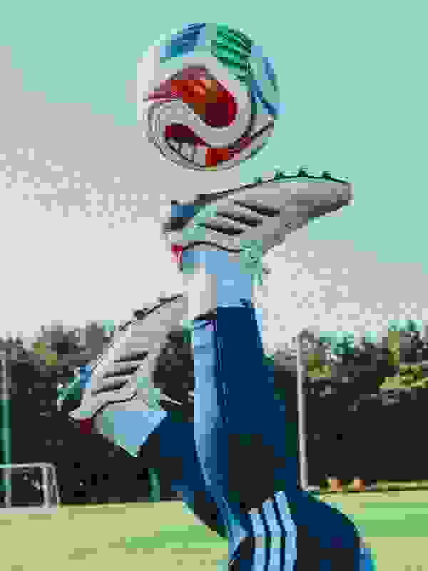 A young person in a blue sports uniform is kicking a soccer ball on a green artificial turf field, with trees and a blue sky visible in the background.