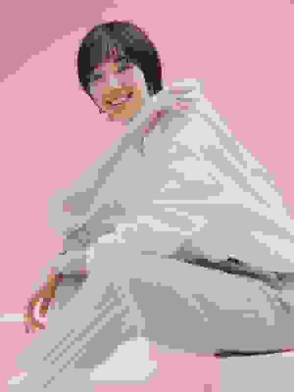 A young man with short dark hair is sitting on a chair, wearing a white shirt and black pants, against a bright blue background.