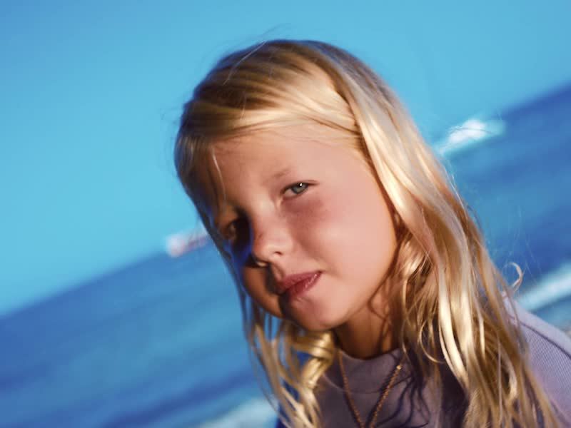 A young girl with long, windblown blonde hair stands against a bright blue sky, wearing a purple sweatshirt with a Disney and Adidas logo.