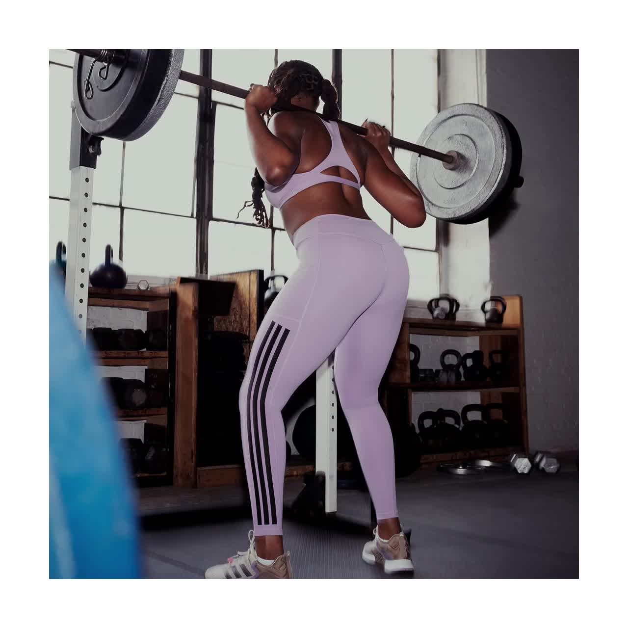 L'immagine mostra una donna in pesi sportivi per il sollevamento pesi in palestra, con varie attrezzature per gli esercizi e scaffali visibili sullo sfondo.