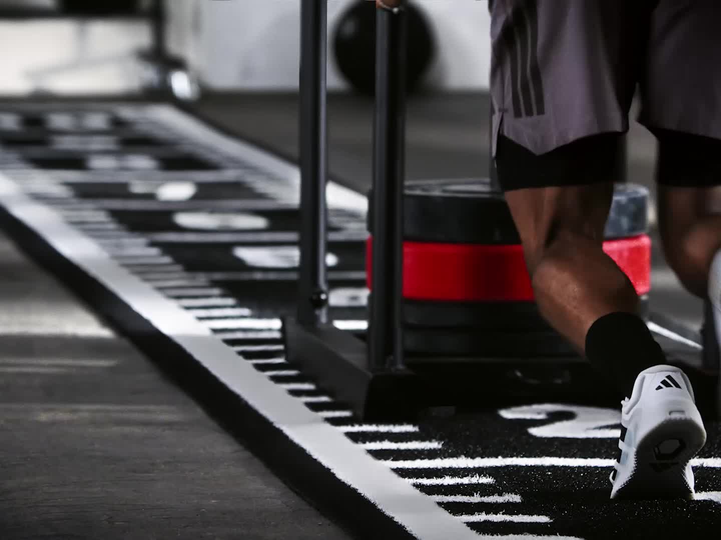 The image shows a person wearing black athletic clothing and white sneakers standing in a gym setting with shelves of equipment in the background.