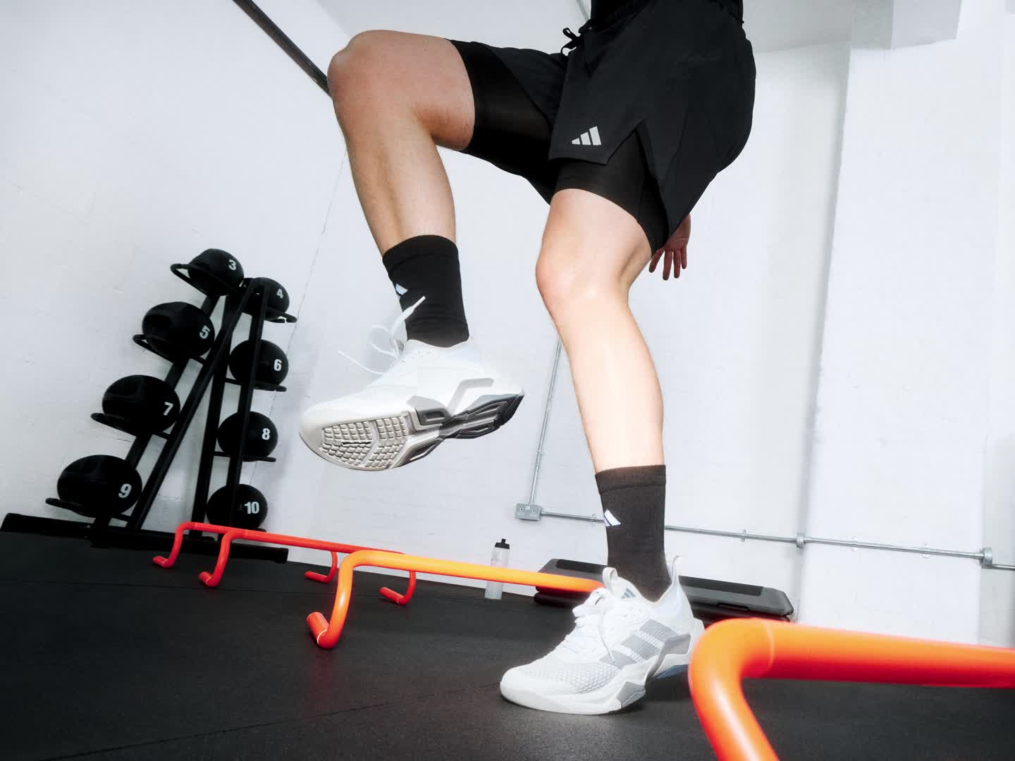 The image depicts a person wearing black athletic clothing and white sneakers, standing on a fitness equipment in a studio setting with various exercise equipment visible in the background.