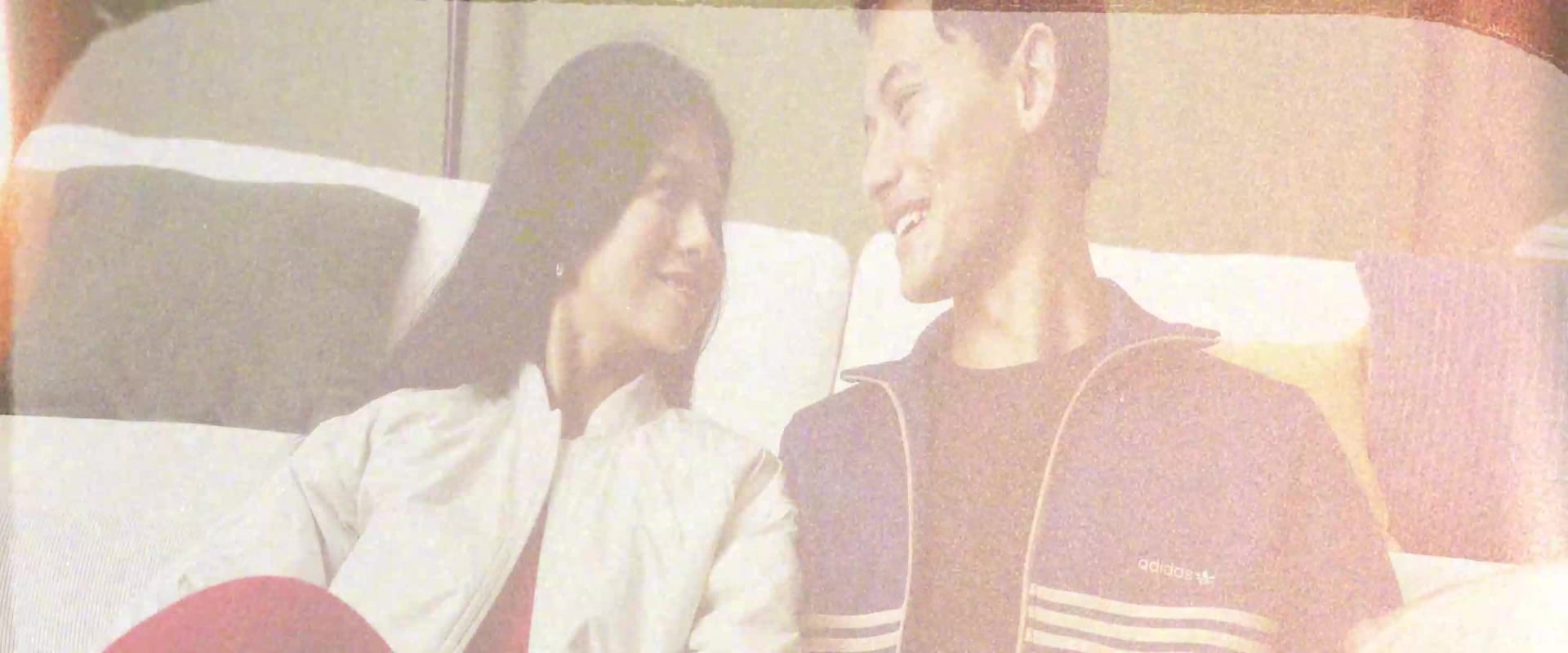 Two individuals, a man and a woman, are seated on a couch in a room with a bookshelf visible in the background.