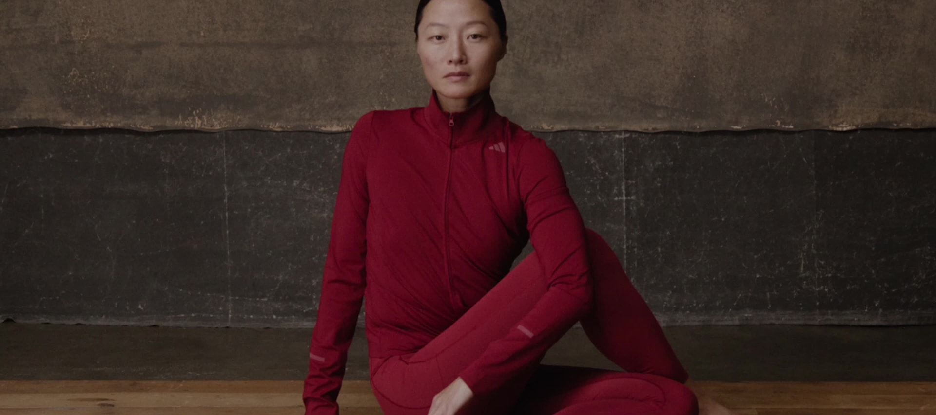 A person in a red leotard performing an acrobatic pose on a wooden floor against a dark background.