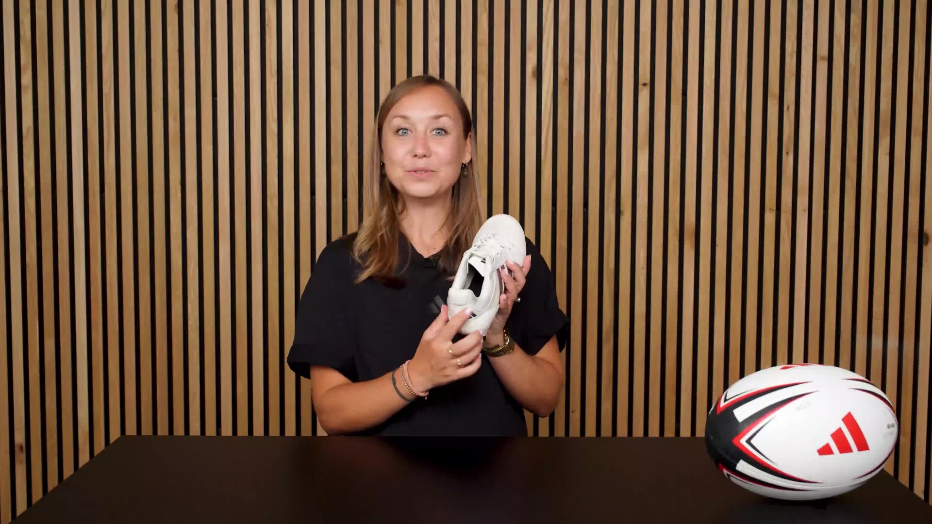 The image shows a woman with long blonde hair wearing a black top, standing in front of a wooden slatted background and holding a pair of white sports shoes.