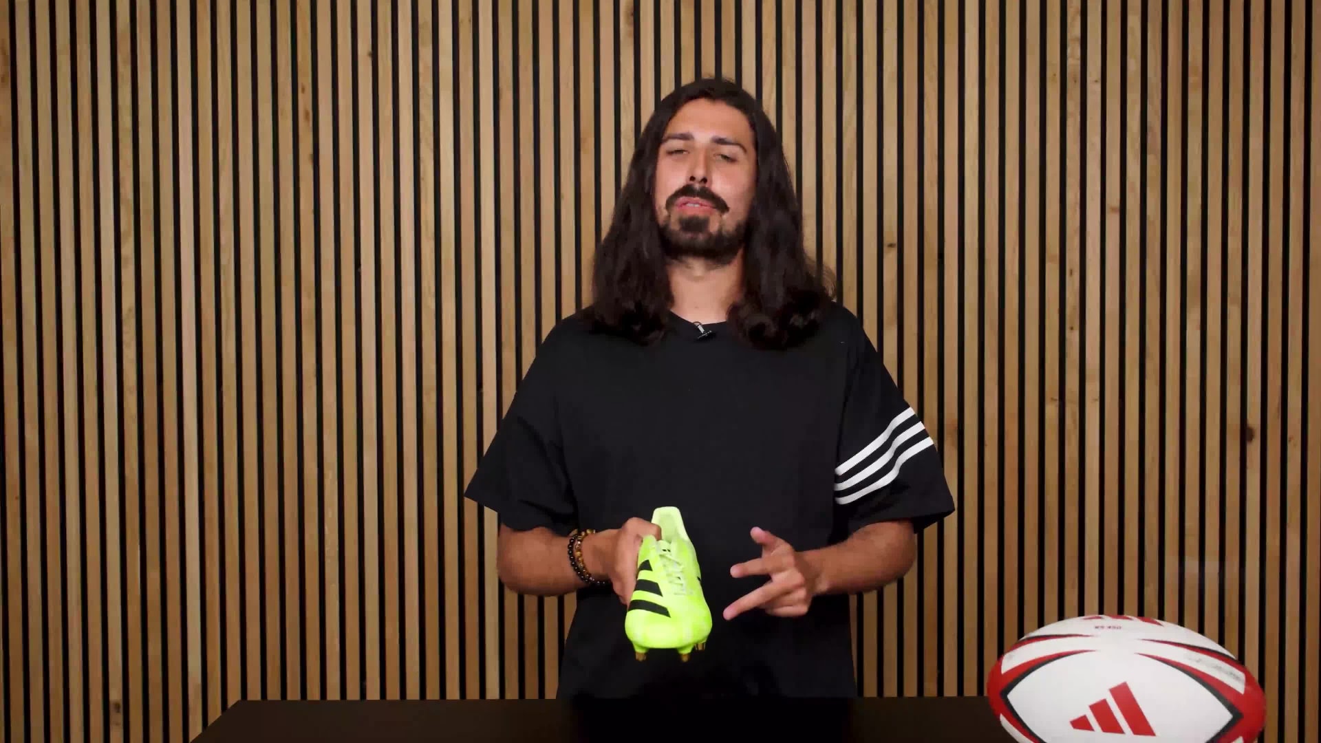 A man with long dark hair and a beard stands in front of a wooden slatted wall, wearing a black shirt with white stripes on the sleeves, and holding a green and white soccer ball.