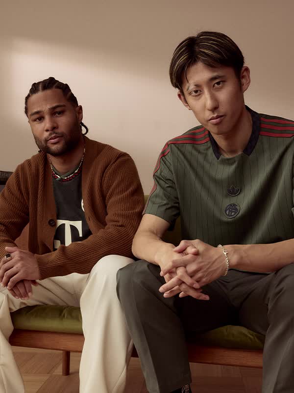 Two individuals wearing sports jerseys, one standing and the other sitting, in a room with a patterned rug on the floor.