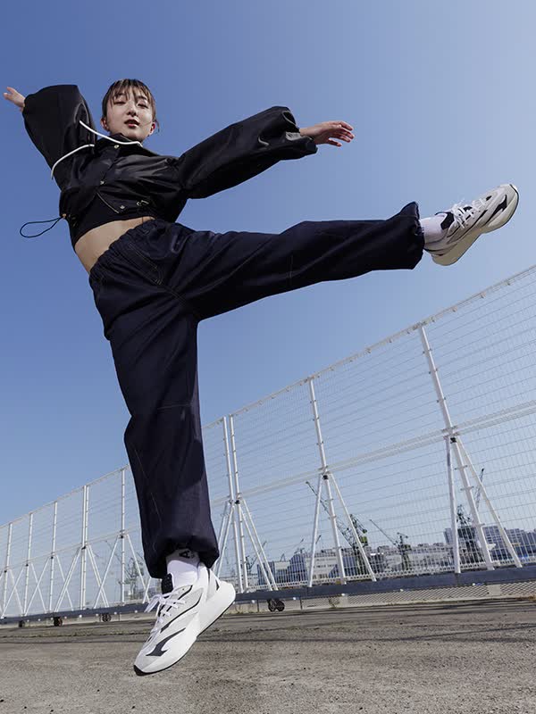 A young woman with long dark hair is captured mid-jump, her body suspended in the air against a backdrop of lush greenery and a clear sky.