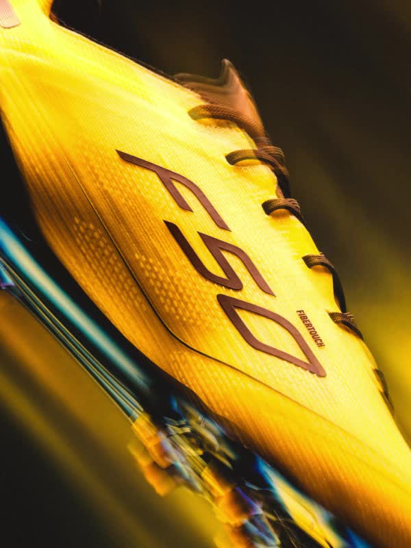 A sleek, black and yellow f50 soccer cleat stands prominently against a vibrant, golden-hued background, casting a dramatic shadow and creating a striking visual contrast.