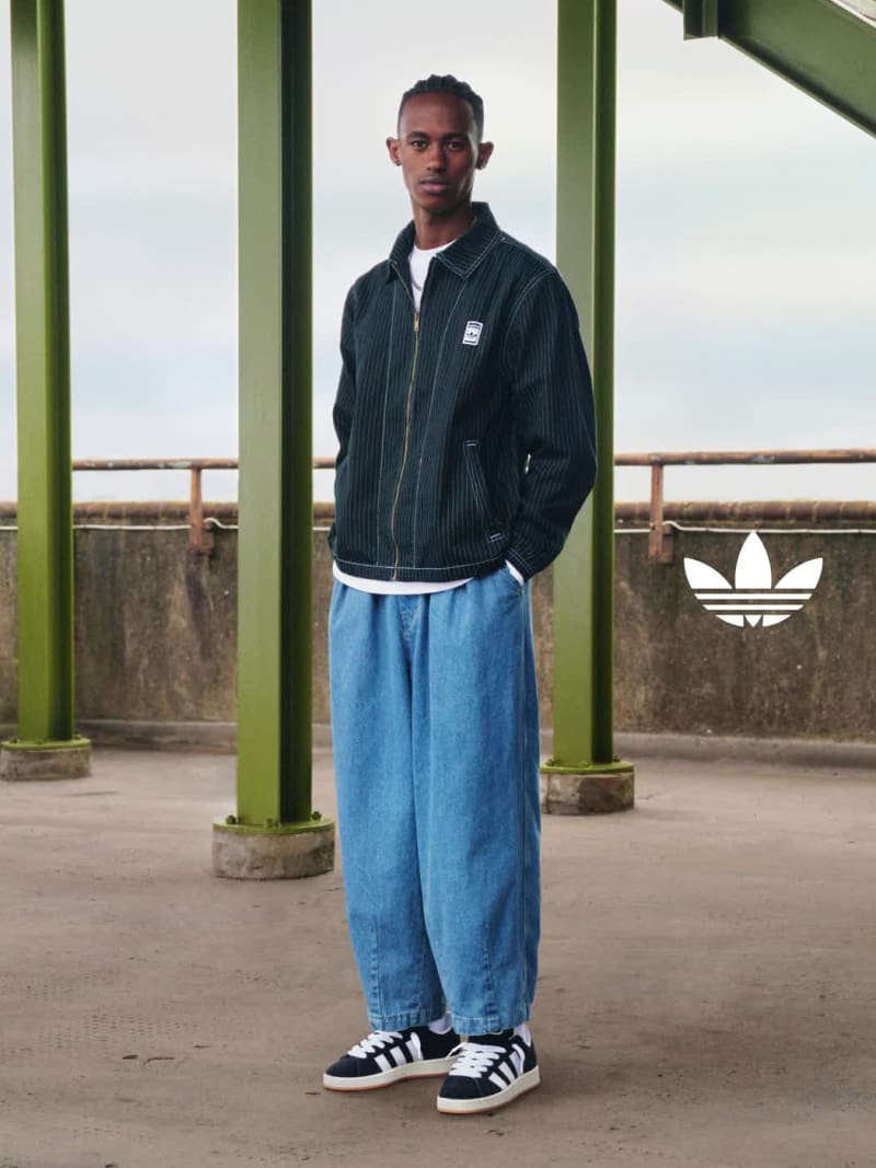 A woman with black braids wears a white and red adidas tee, a long beige skirt, and black adidas Campus 00s shoes on a concrete street.