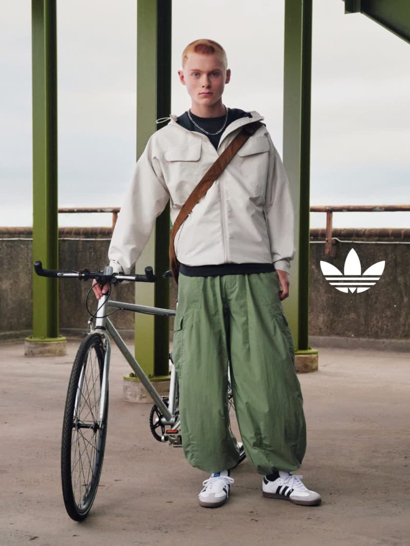  A girl with black braids poses confidently in a white adidas top, blue jeans, and black adidas Samba shoes on a concrete street. 