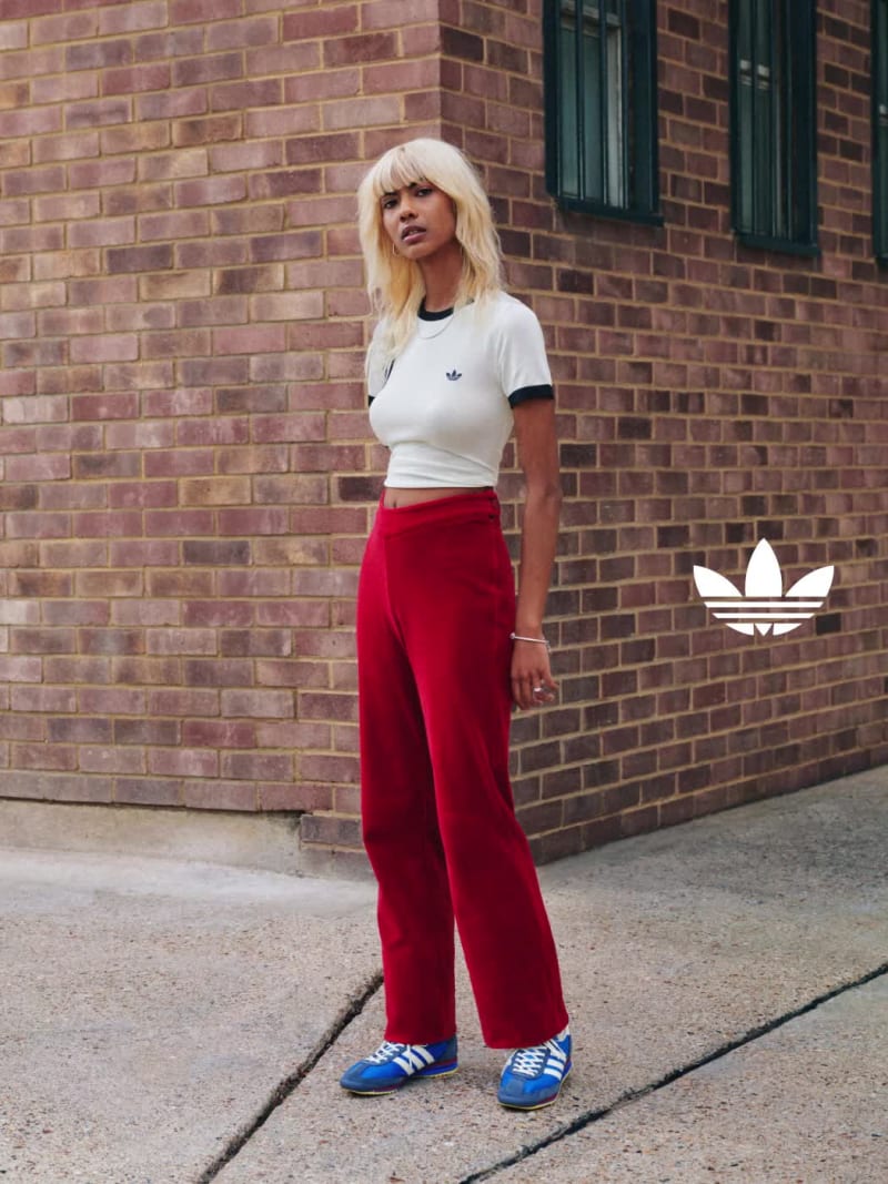 A man and a woman strike a pose in front of a brick wall, wearing adidas apparel and blue adidas SL 72 sneakers.