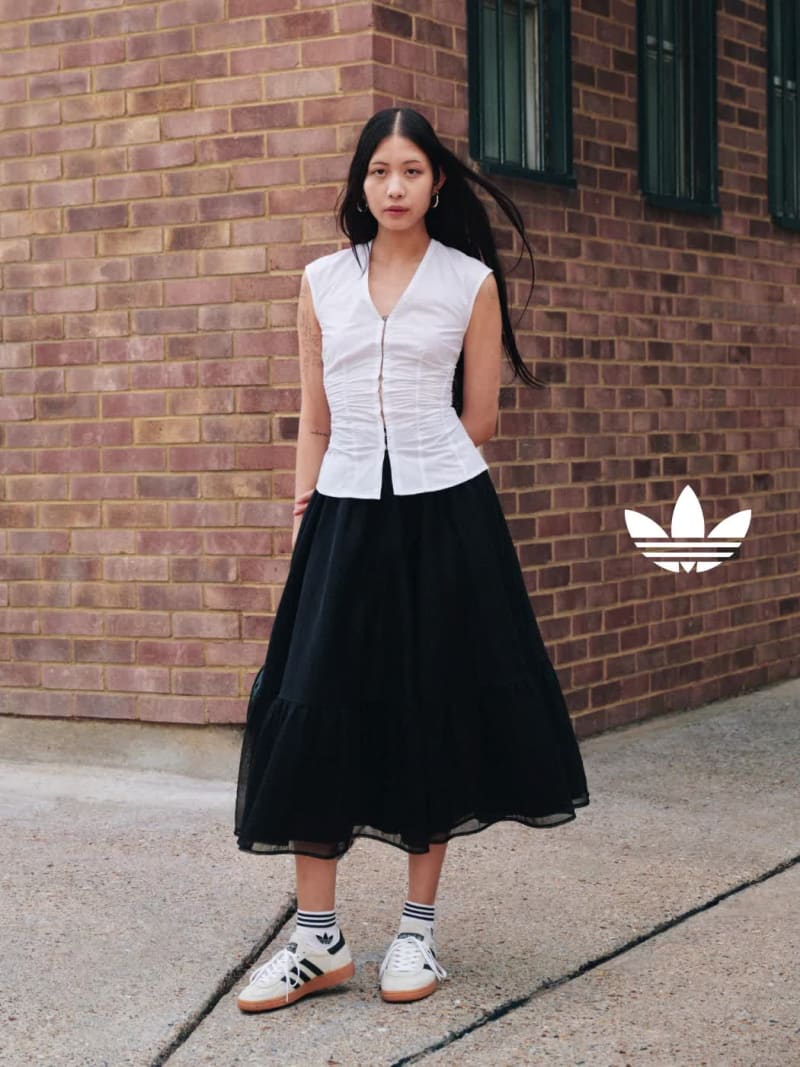 A man and a woman in adidas apparel and white and black adidas Handball Spezial shoes stand in front of a brick wall in the daytime.
