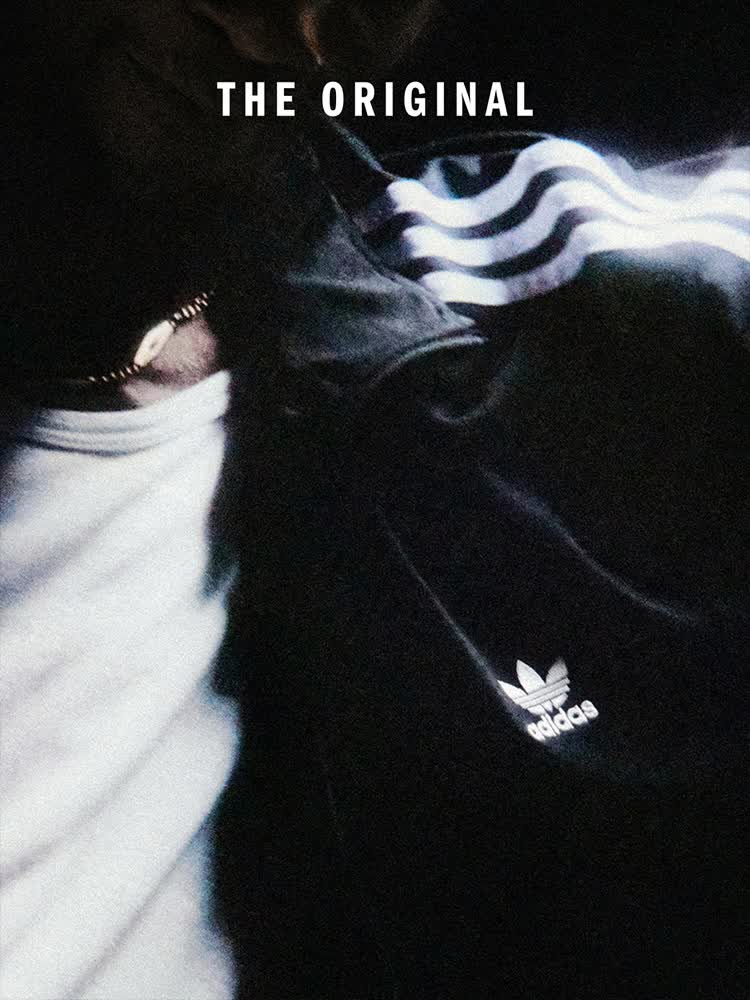 Person in a black Adidas Firebird jacket stands with arms outstretched in a crowded space; Firebird and Adidas logo are visible against a dark background.