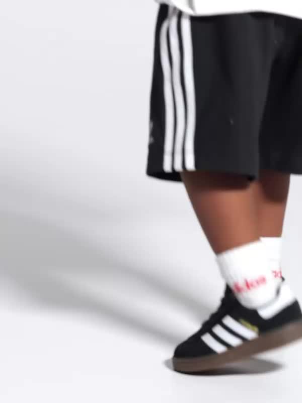 A young child with curly hair wearing a white shirt and black shorts is sitting on a white surface against a plain white background.