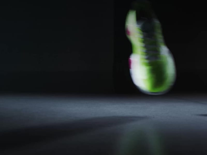 A vibrant green and pink sneaker rests on a dark surface, casting a dramatic shadow against the black background.